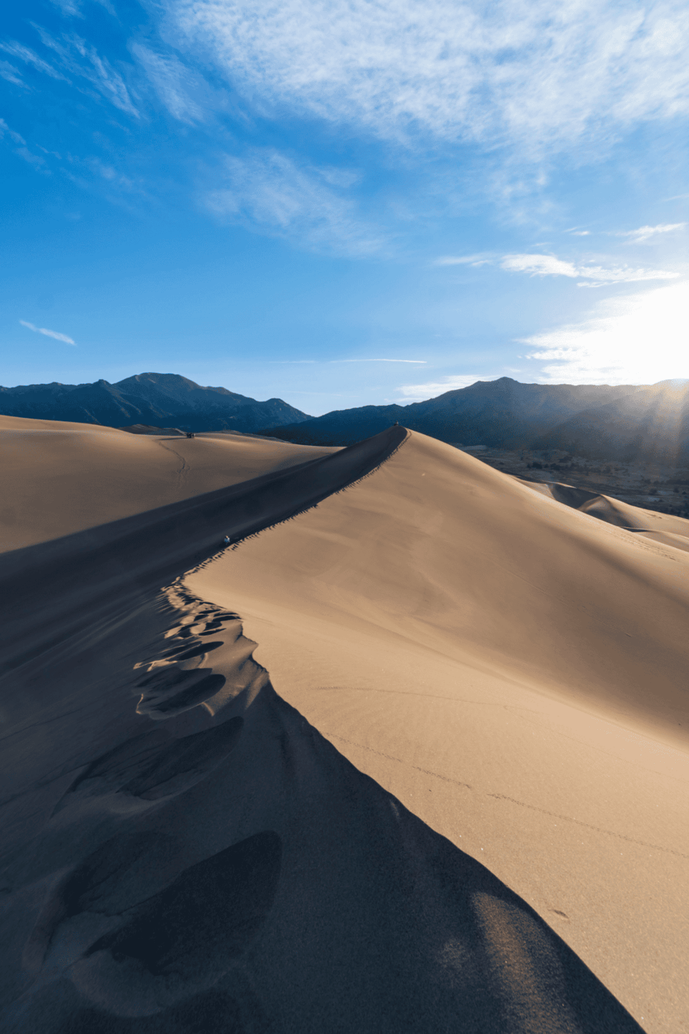 Sweeping desert dunes with mountain backdrop under blue sky, ideal for adventure and outdoor exploration.