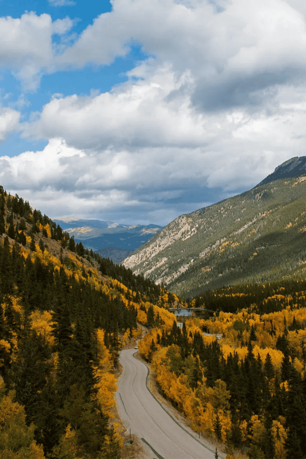 Winding mountain road through fall foliage with scenic valley and blue sky in background.