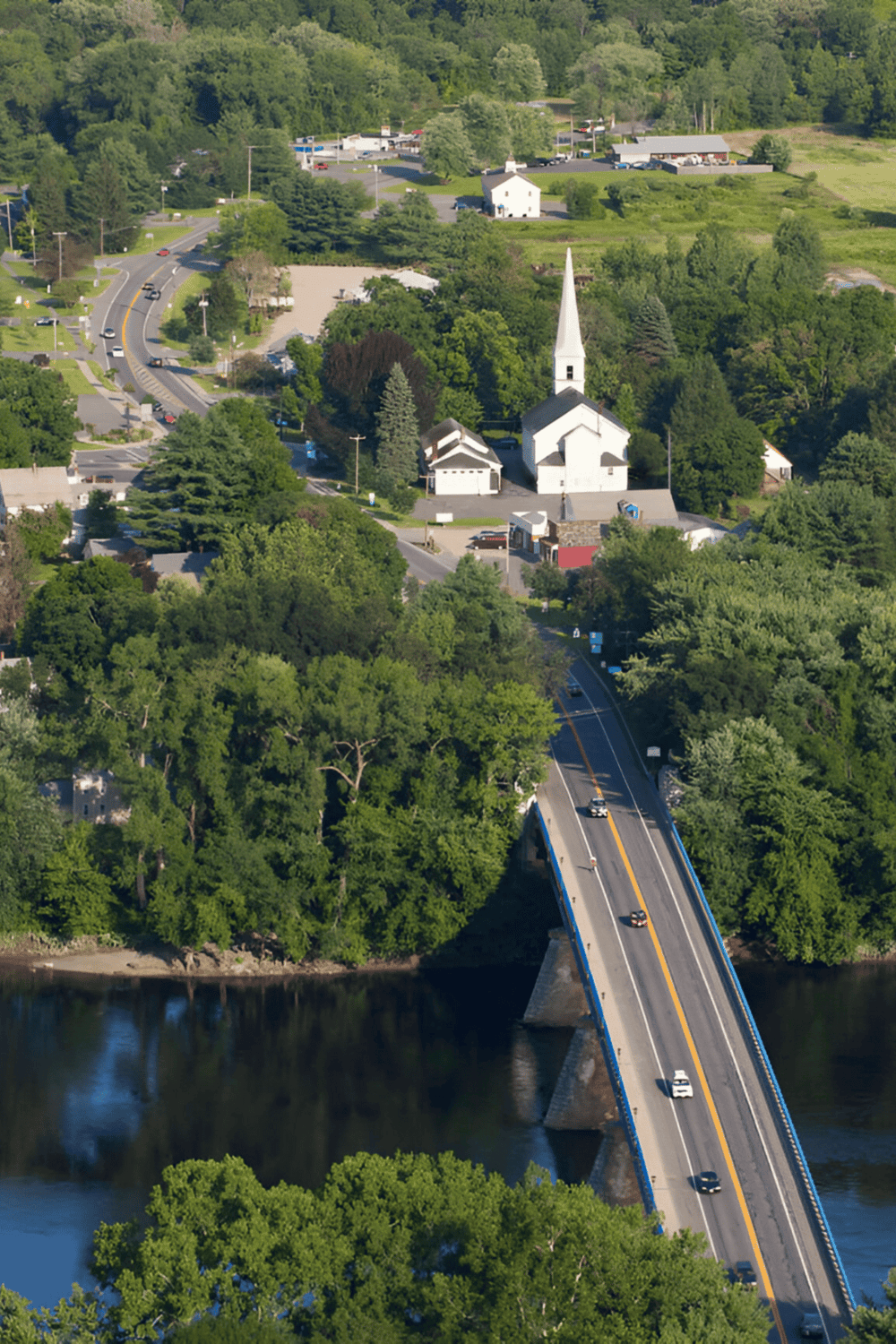 Aerial view of a charming small town with a white church, lush greenery, and a bridge over a river.