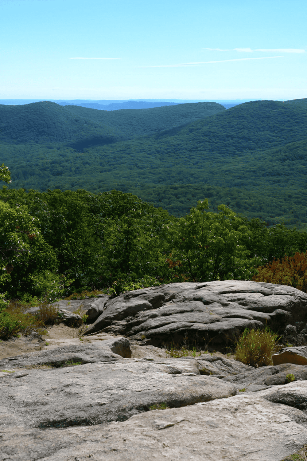 Vast green mountain landscape with rocky foreground, lush trees, and blue sky, showing outdoor scenic nature views.
