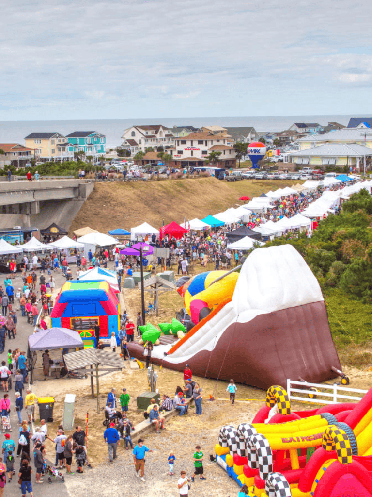 Vibrant outdoor fair featuring colorful inflatable bounce houses, food stalls, and crowded gatherings at a seaside community.