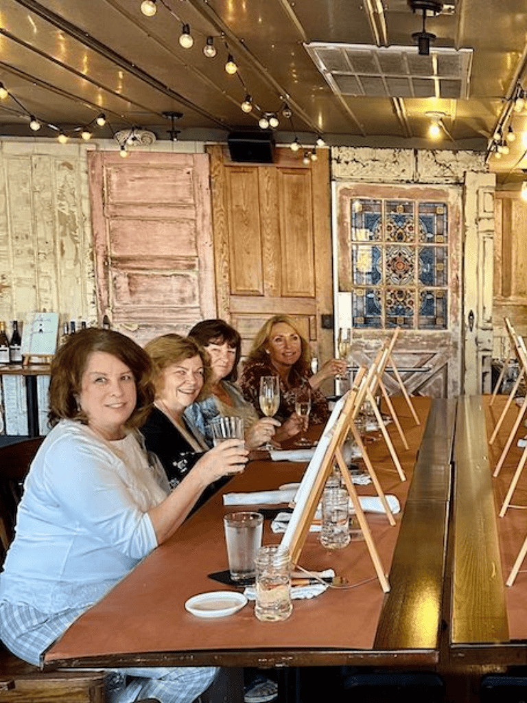 Elegant women participating in a paint and sip wine tasting event at a rustic-themed venue.