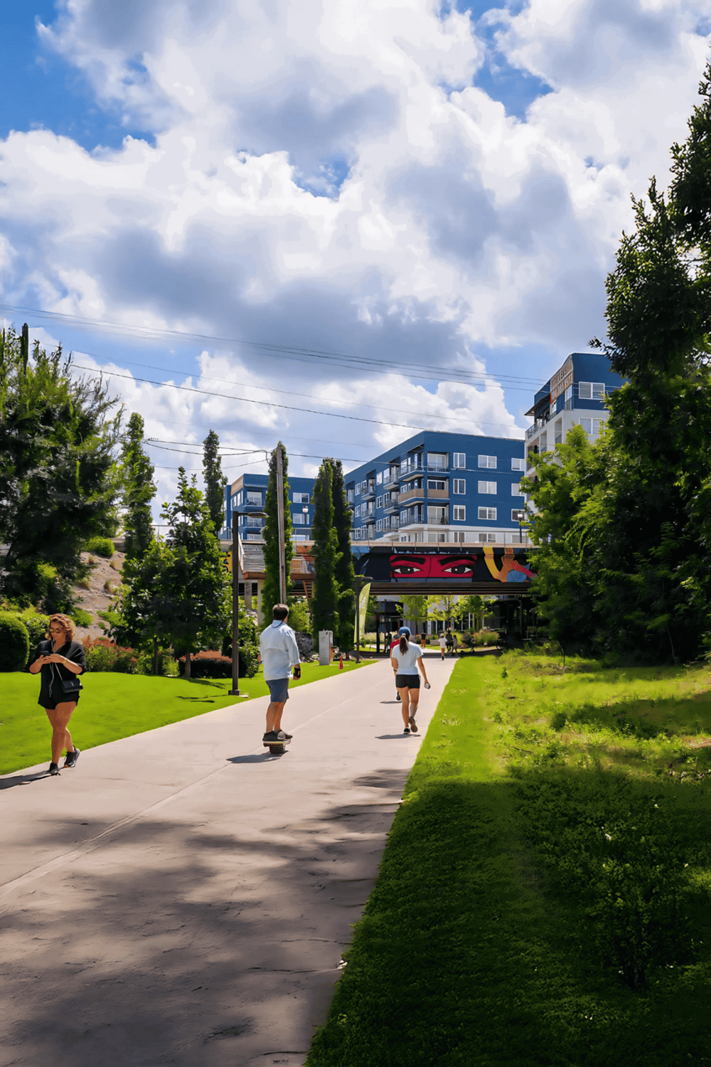 Modern urban park with people skating and walking, surrounded by trees, apartment buildings, and a blue sky.
