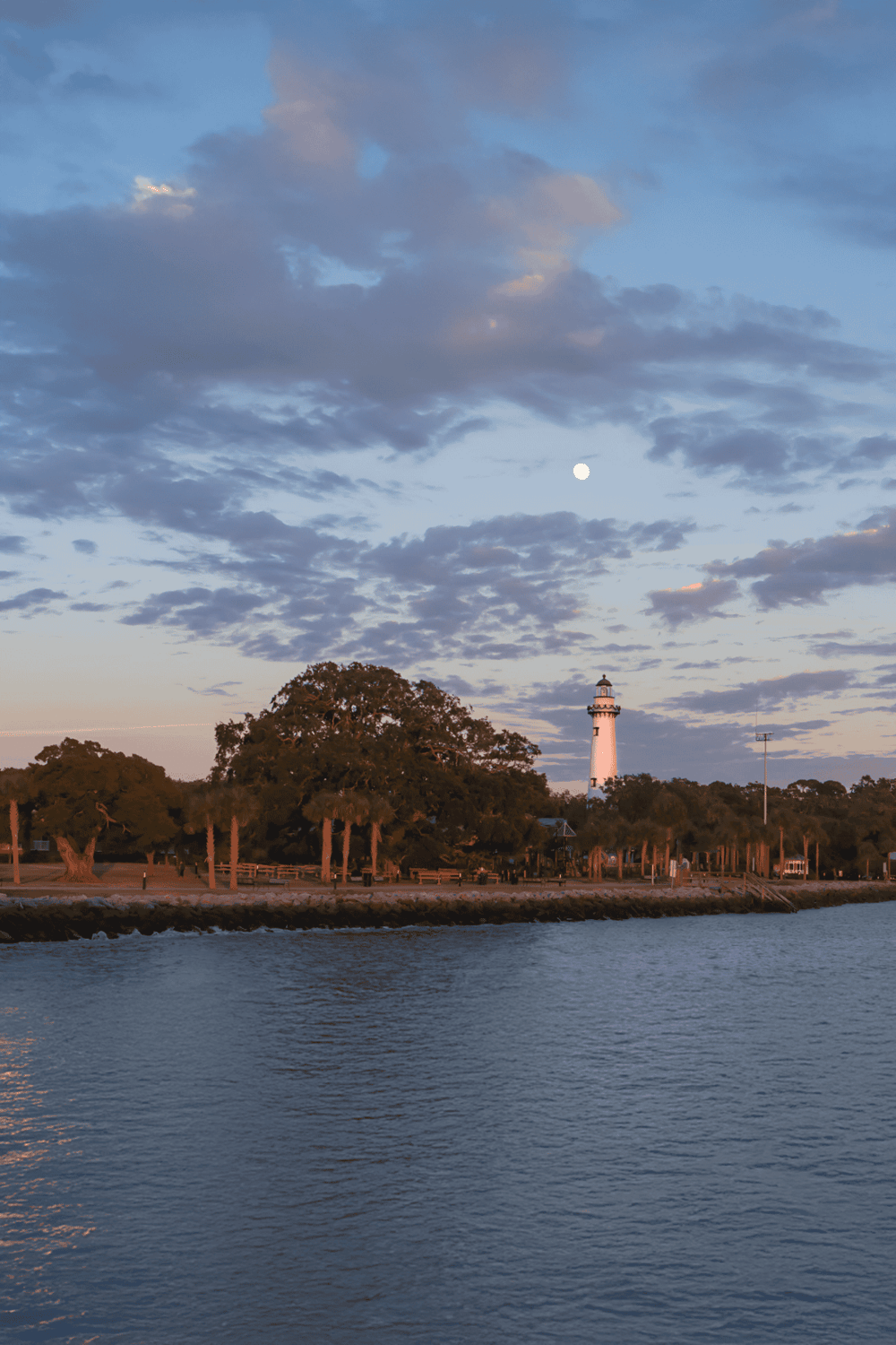 A serene scene of the lighthouse at dusk with a full moon, trees, and calm water, showcasing scenic coastal navigation tools.
