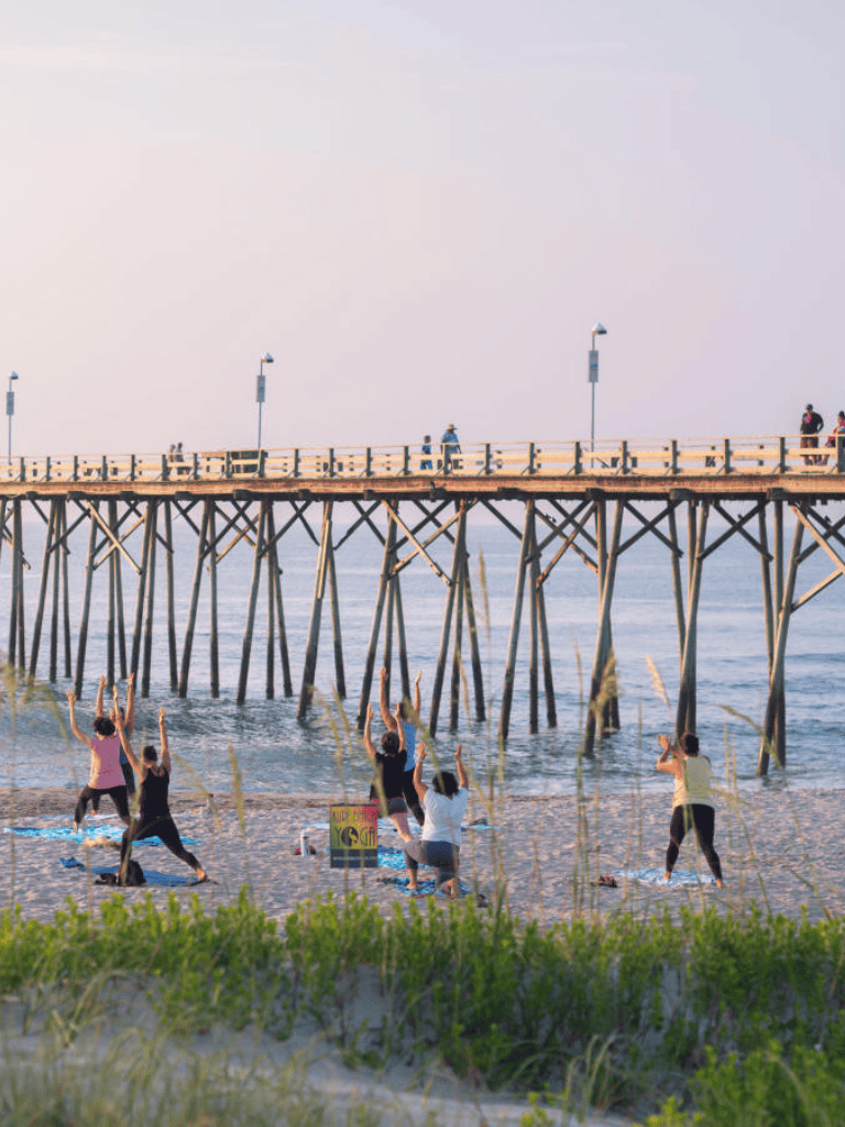 People doing yoga on the beach near a wooden pier during sunset, promoting wellness and outdoor fitness.
