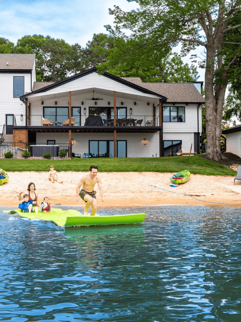 Bright lakeside house with a sandy beach, family enjoying paddleboarding, perfect for outdoor recreation.