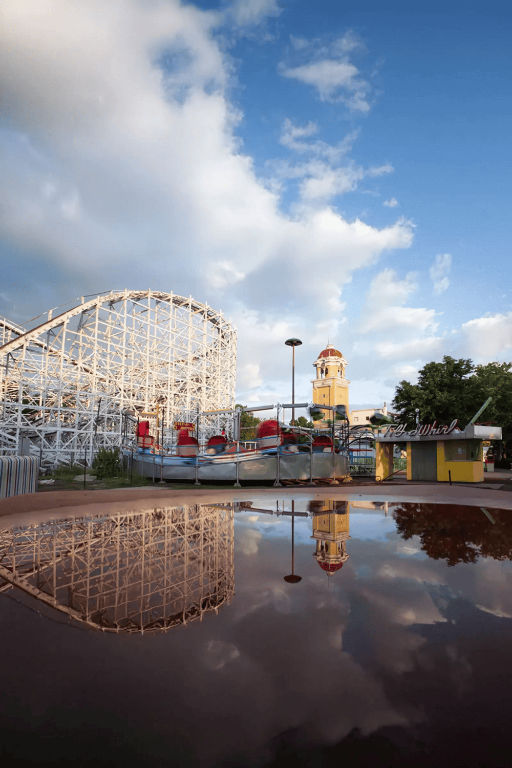 Colorful amusement park rides with a wooden roller coaster, blue sky, and reflection in water, offering family-friendly entertainment.