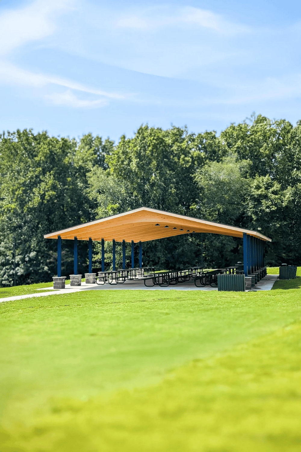 Modern outdoor picnic shelter with wooden roof and picnic tables in a lush park setting.