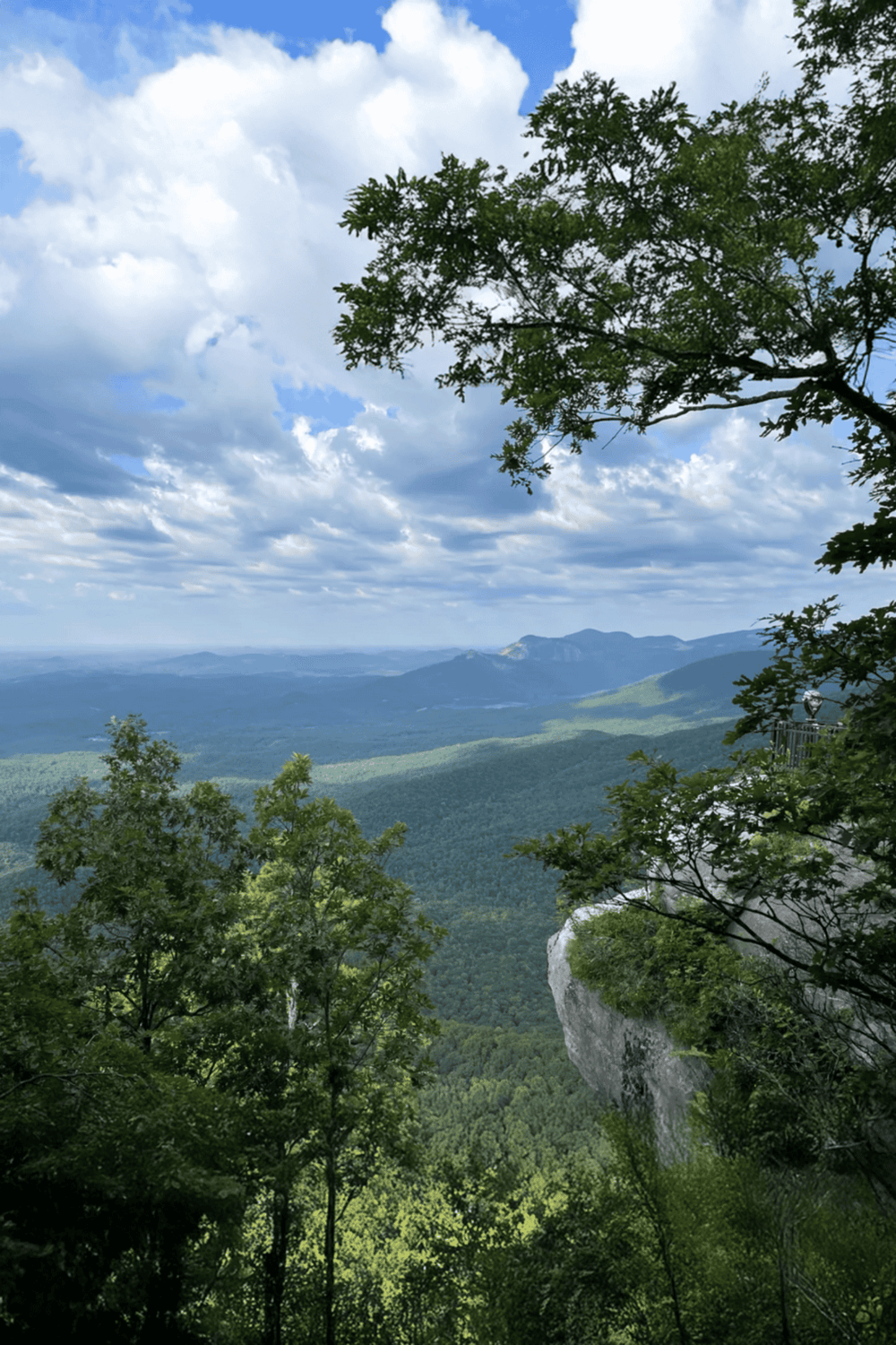 Vast mountain landscape with lush green trees, cloudy sky, and distant ridges in the background.