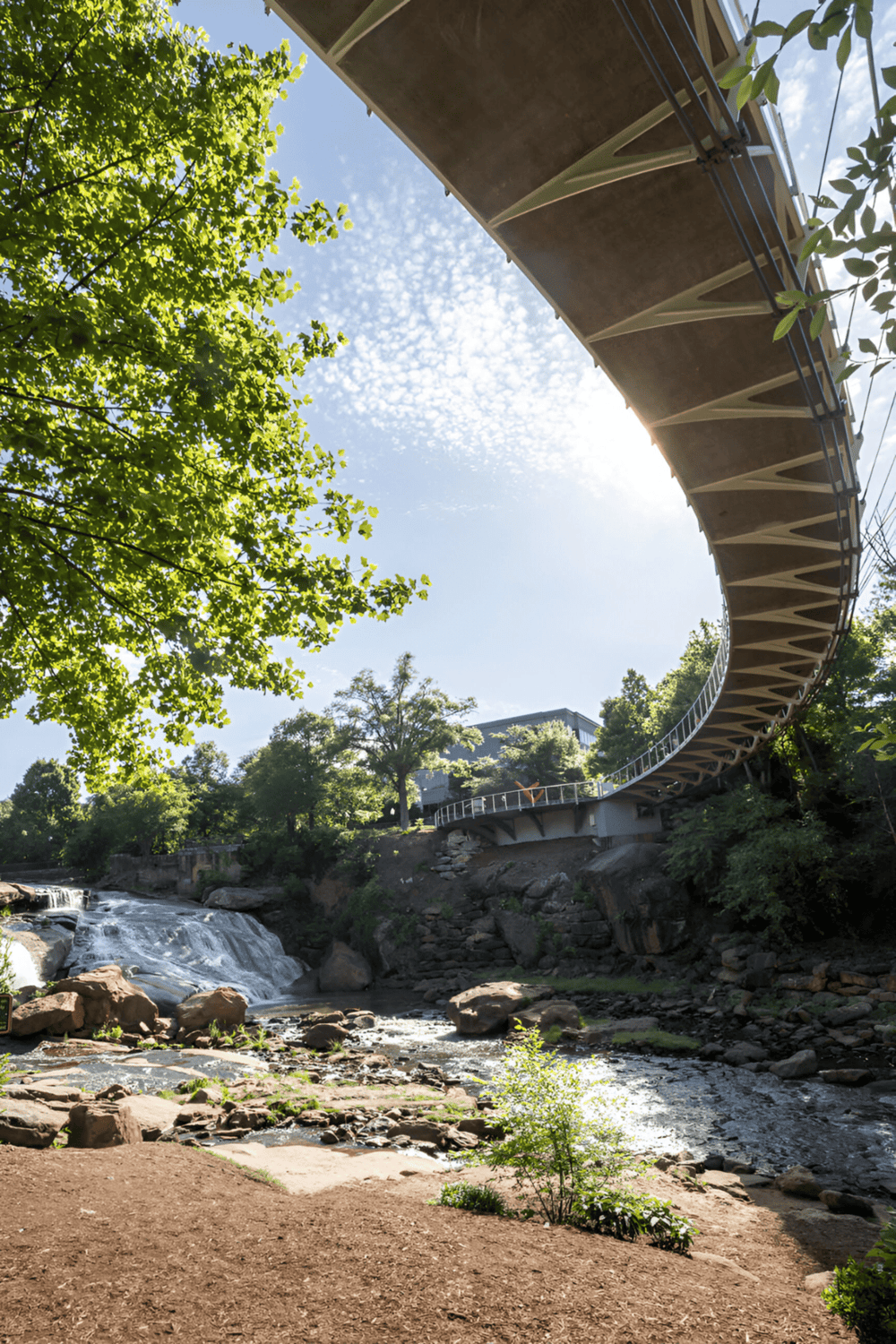 Suspended pedestrian bridge over river with lush green trees and blue sky, modern architecture, scenic outdoor view.