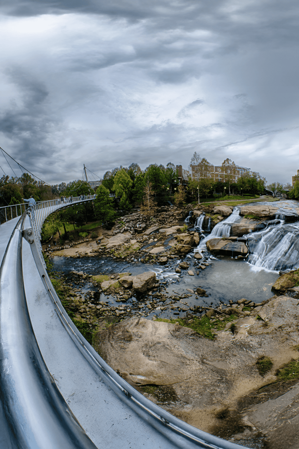 Scenic view of a city park with a waterfall, bridge, and lush greenery under a cloudy sky.