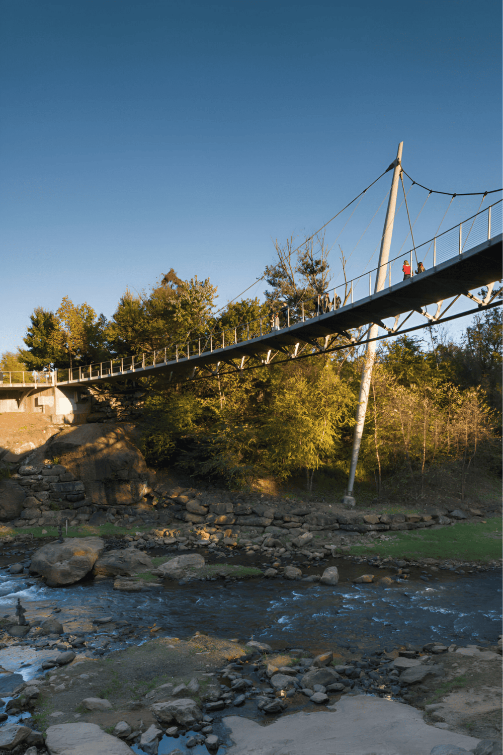 Suspension bridge over rocky river with lush trees and clear blue sky in background.