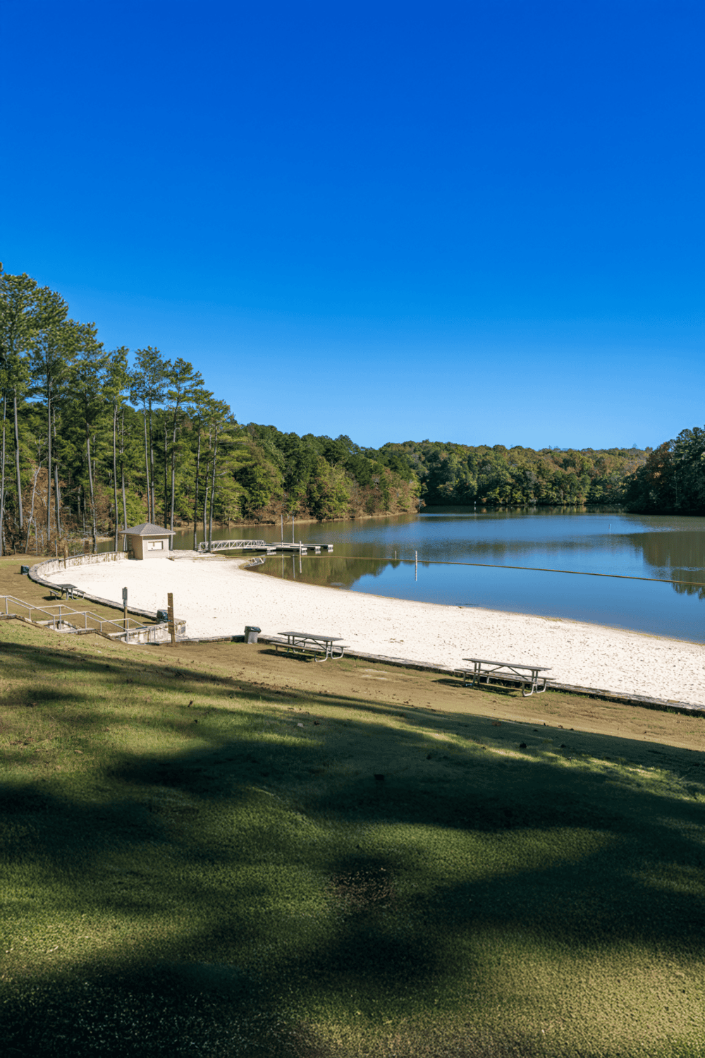 Quiet lakeside park with sandy beach, boat docks, and lush trees under clear blue sky.