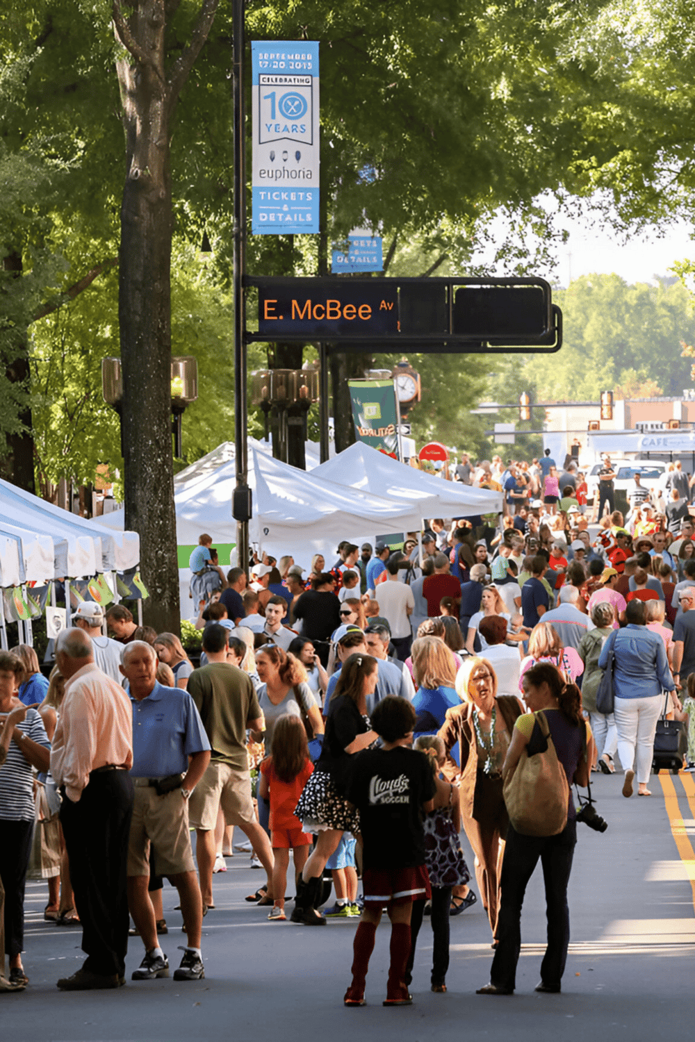 Crowd at outdoor event with tents on a tree-lined street, celebrating arts festival and local community.