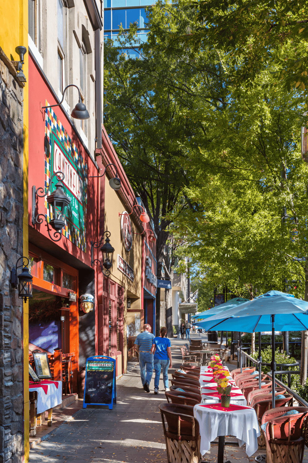 Colorful street scene with outdoor restaurant tables and greenery in a vibrant downtown area.