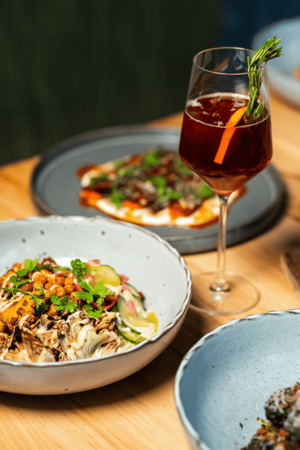 A refreshing drink with herbs served alongside gourmet salads and pizza on a wooden table in a modern restaurant.