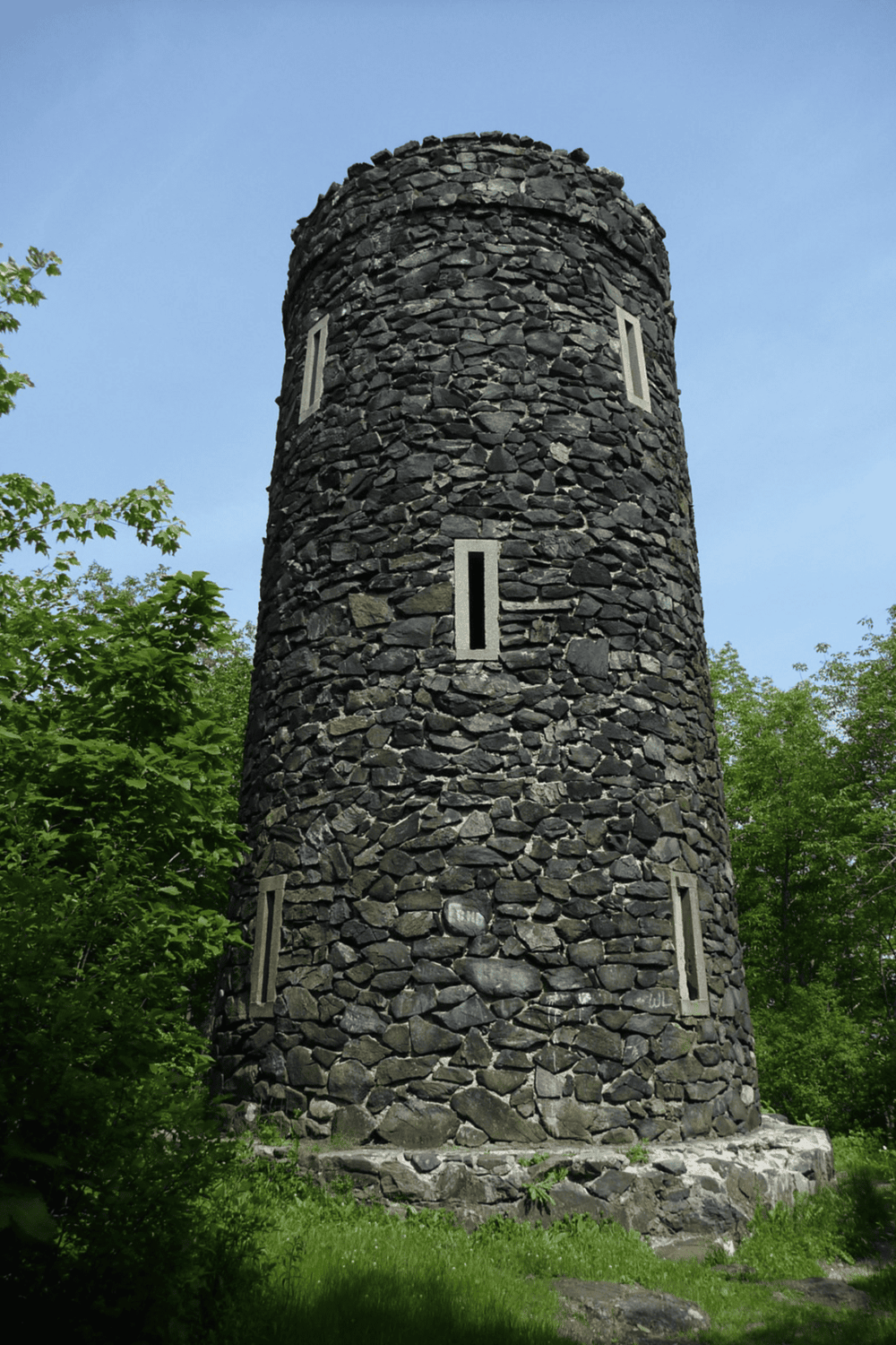 Weathered stone tower with narrow vertical windows, surrounded by lush greenery.