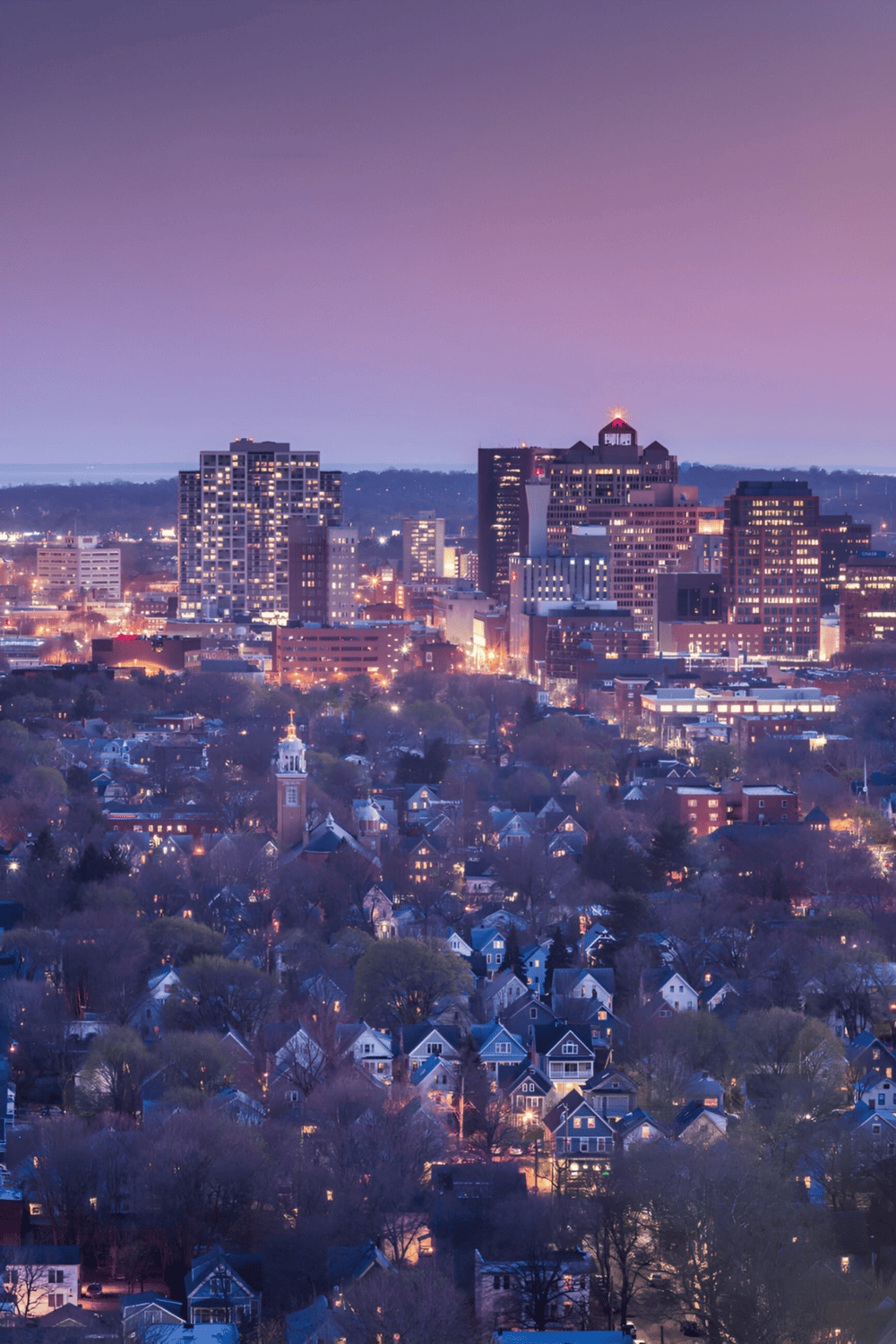 Bright city skyline at dusk with illuminated buildings and residential neighborhood, showcasing urban and suburban landscapes.