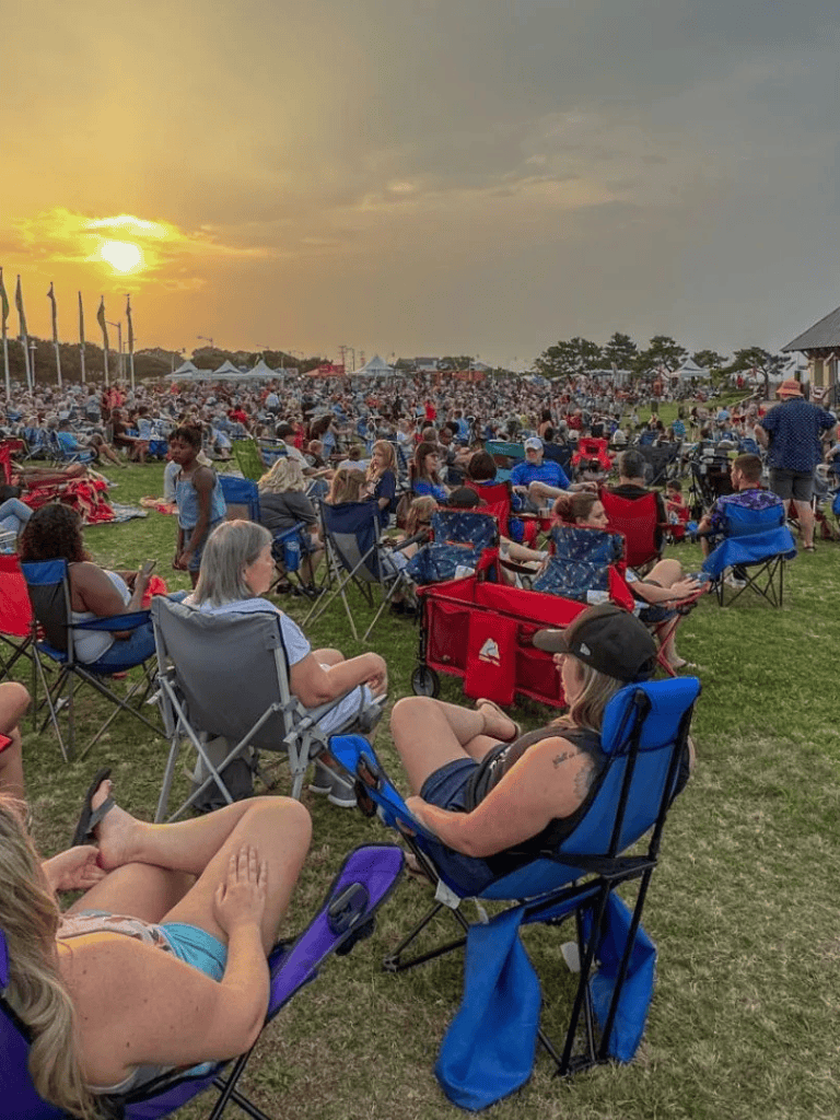 Sunset outdoor concert with large crowd, seating in lawn chairs, community event, festival atmosphere, family fun, summer evening.