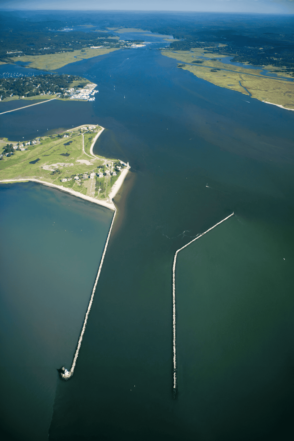 Aerial view of a coastal area featuring a marina, breakwaters, and lush green landscapes.