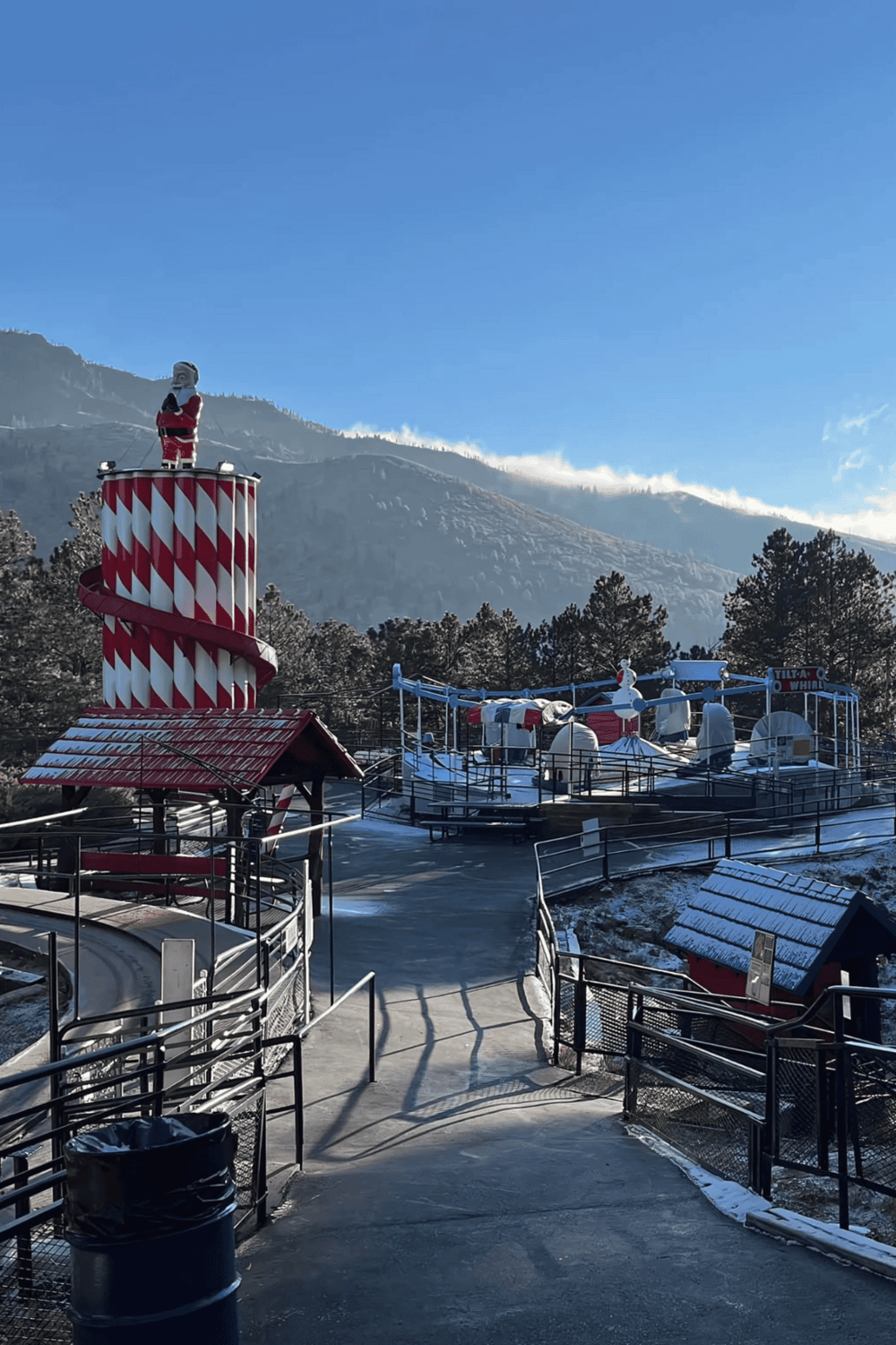 Santa-themed amusement park with a snow-covered carousel and a red-and-white striped lighthouse in a mountain setting.