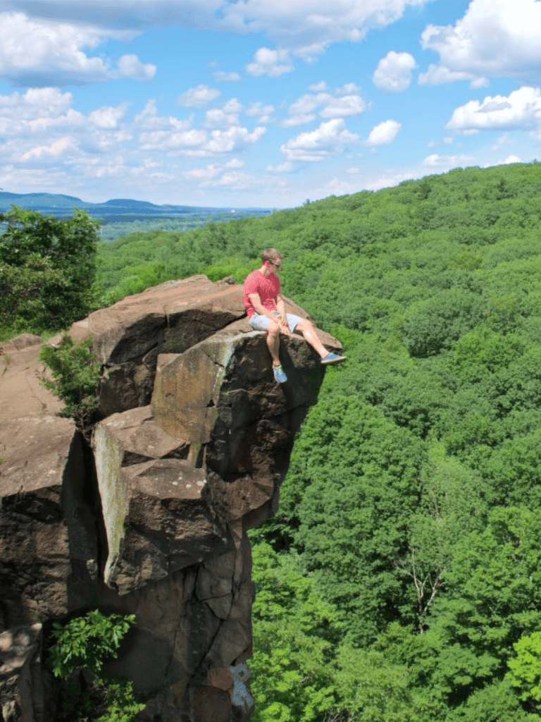 Cliff lookout with a person sitting on rocks above lush green forest in a scenic landscape.