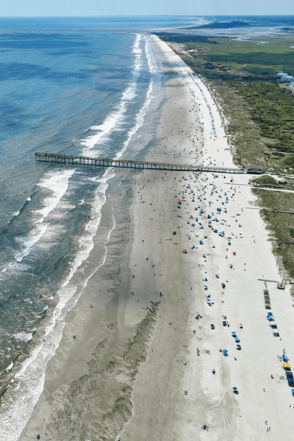 Aerial view of a scenic beach with pier, ocean waves, and colorful umbrellas.