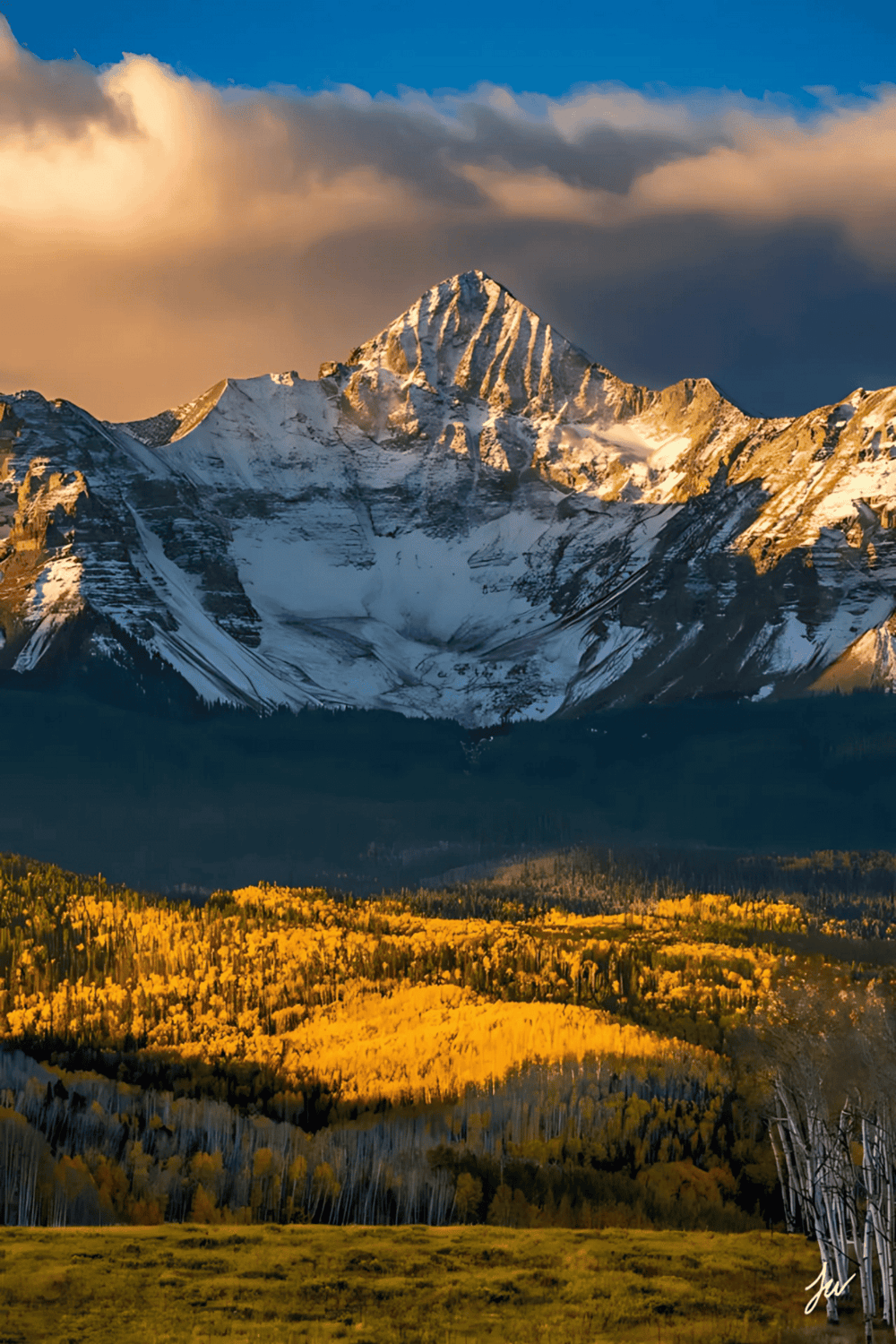 Majestic snow-capped mountain peak with vibrant autumnal forest and cloudy sky in the background.
