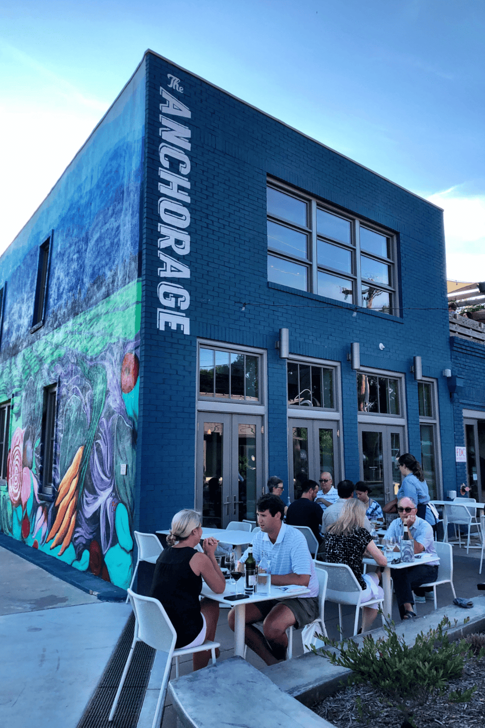 Modern blue brick building with outdoor dining at Quest For Directions restaurant, vibrant mural wall, people enjoying meals.