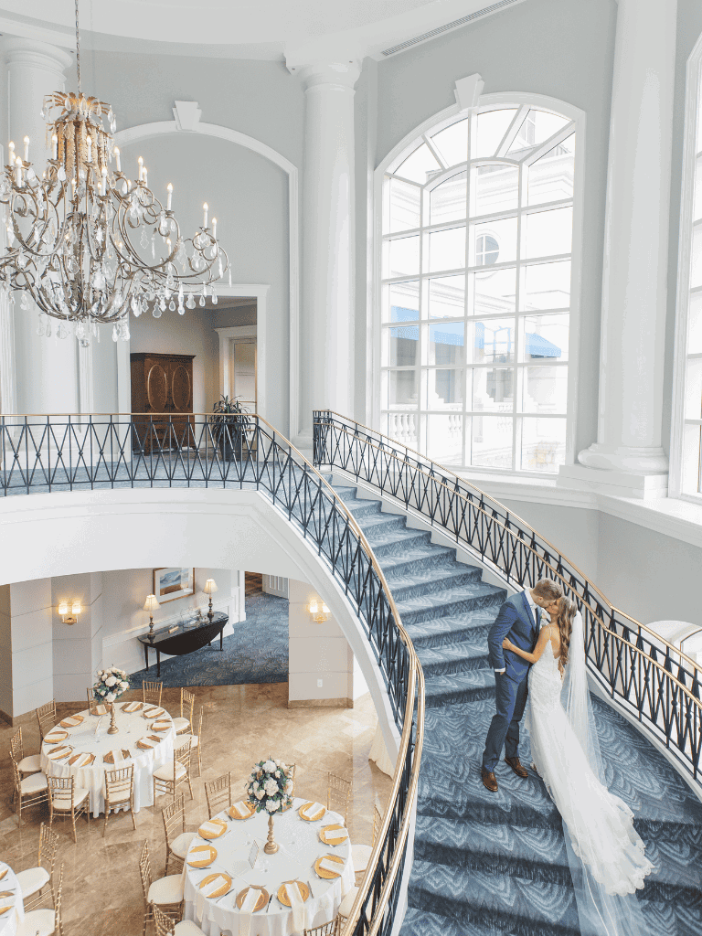 Elegant wedding couple on staircase in grand ballroom, soft natural lighting, luxury venue, romantic event venue, upscale wedding location.