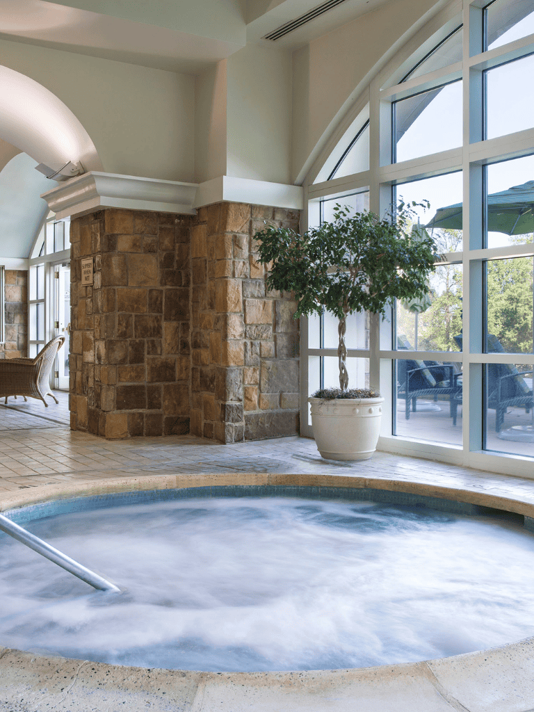 Relaxing indoor hot tub with large windows and natural stone decor for health and wellness.
