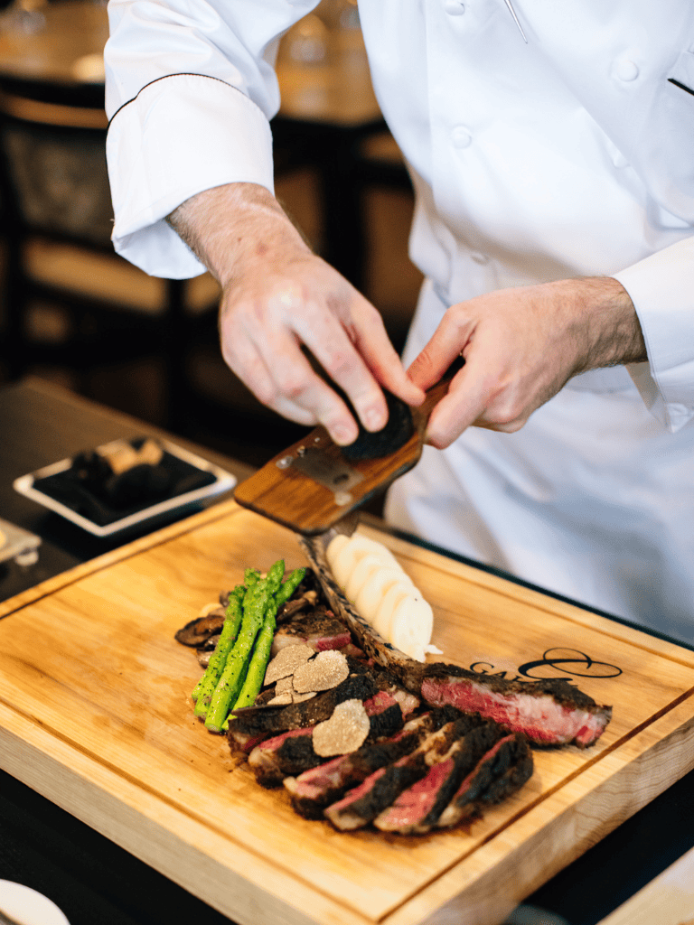 Prime-grade steak being expertly sliced with truffle shavings and fresh vegetables on a wooden serving board.