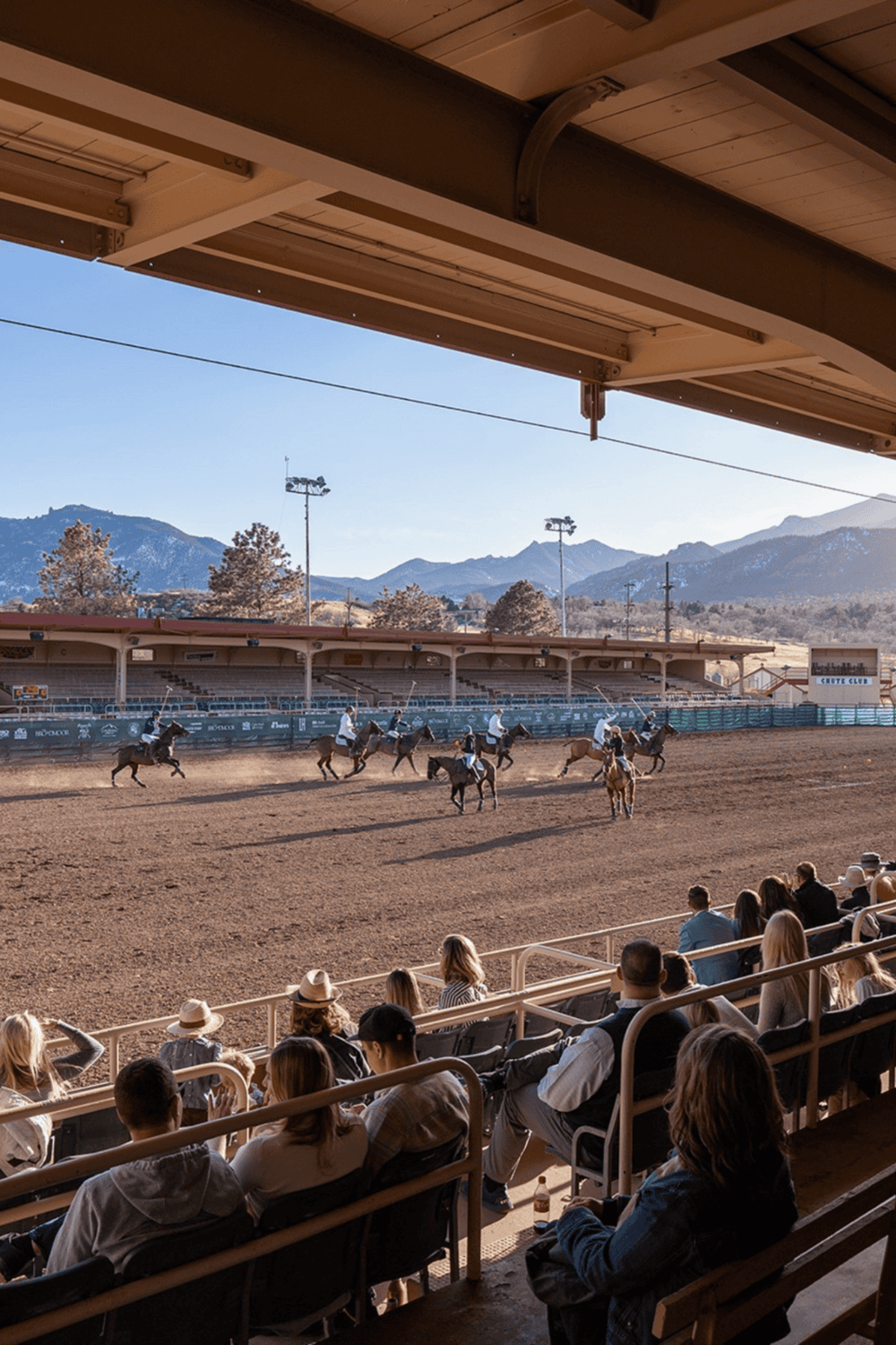 Horseback riding event at an outdoor arena with spectators, mountains in the background, sunny weather, and stadium seating.