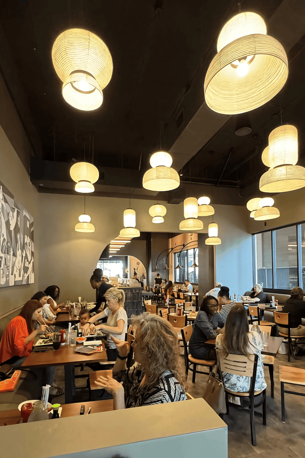 Cozy modern restaurant interior with warm hanging pendant lights, diverse group dining, and large windows for natural light.