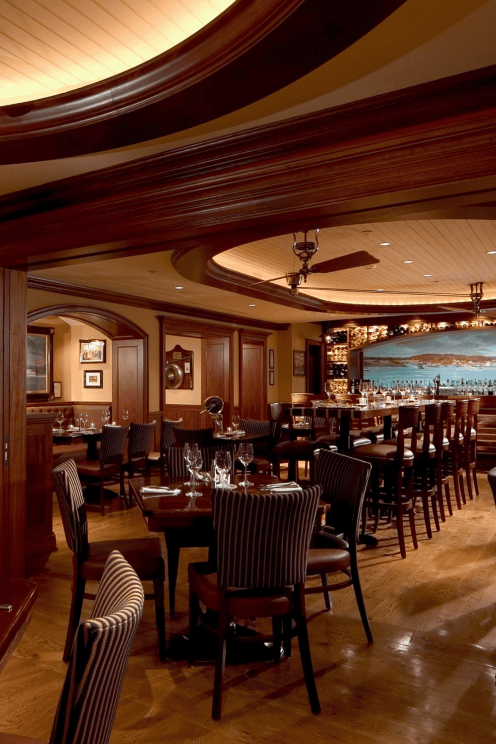 Cozy upscale restaurant interior with wooden accents, elegant table settings, and a waterfront view in the background.