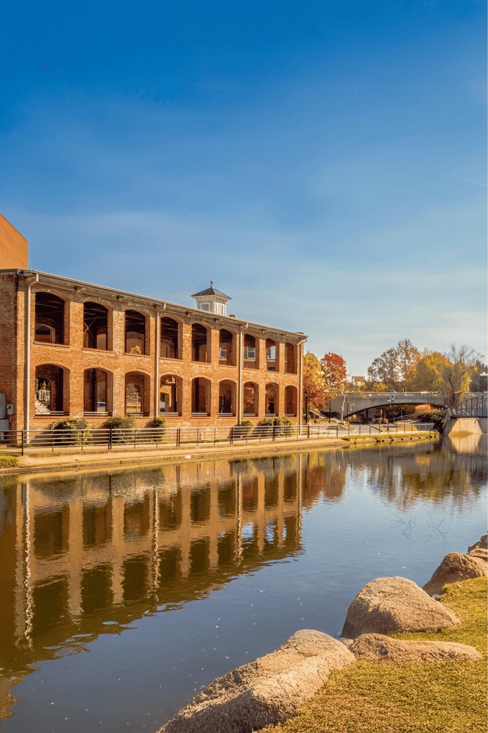 Historic brick building along a peaceful river with autumn foliage and a clear blue sky.