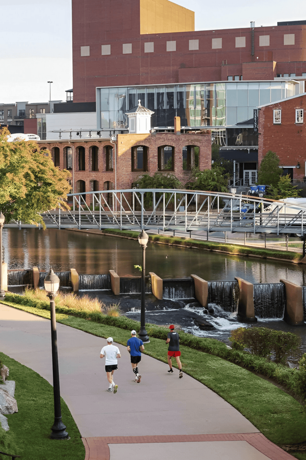 Modern urban park with runners, waterfall, historic brick buildings, and city skyline in the background.