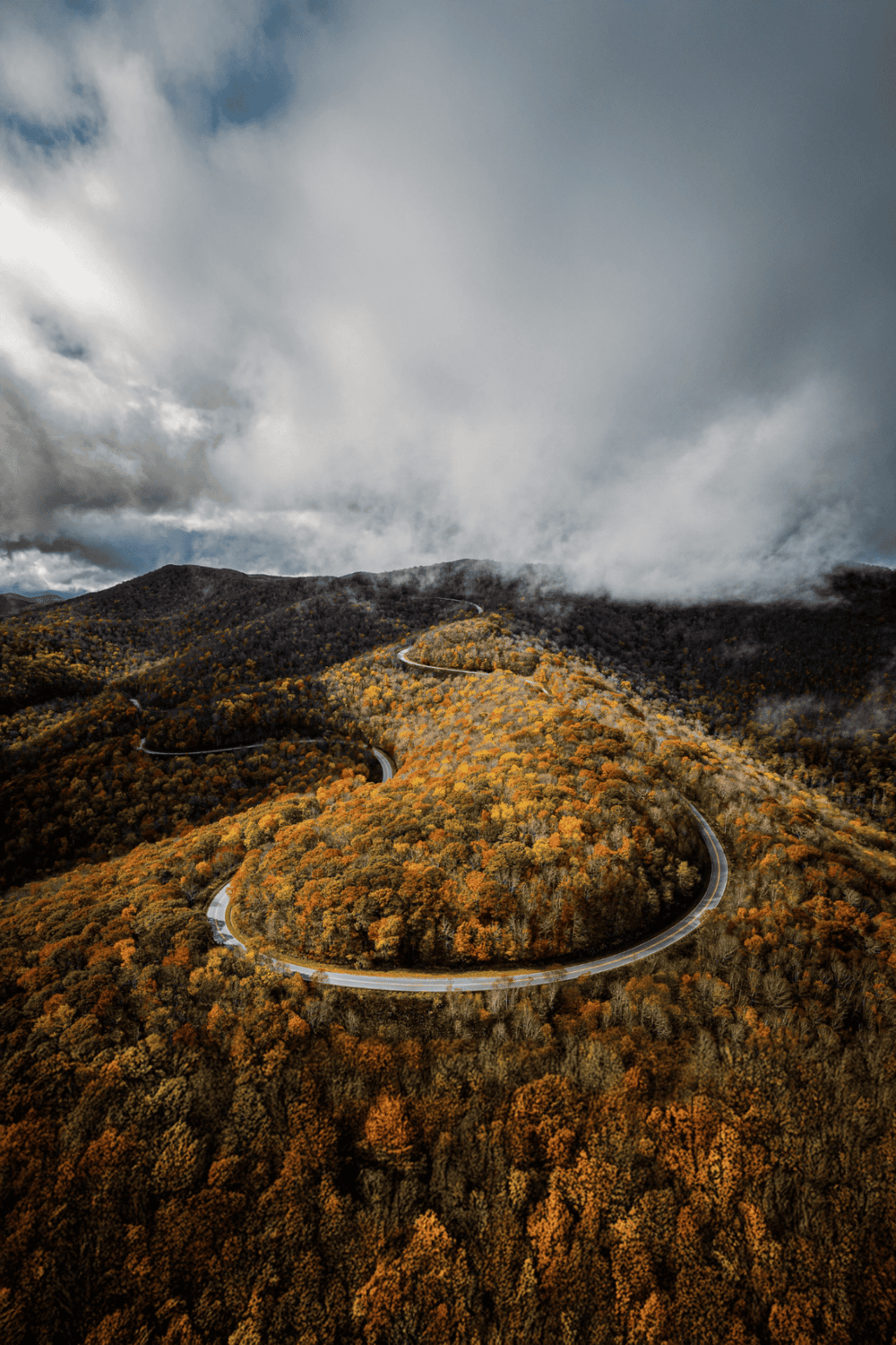 Winding mountain road through fall foliage with cloudy sky in the background.