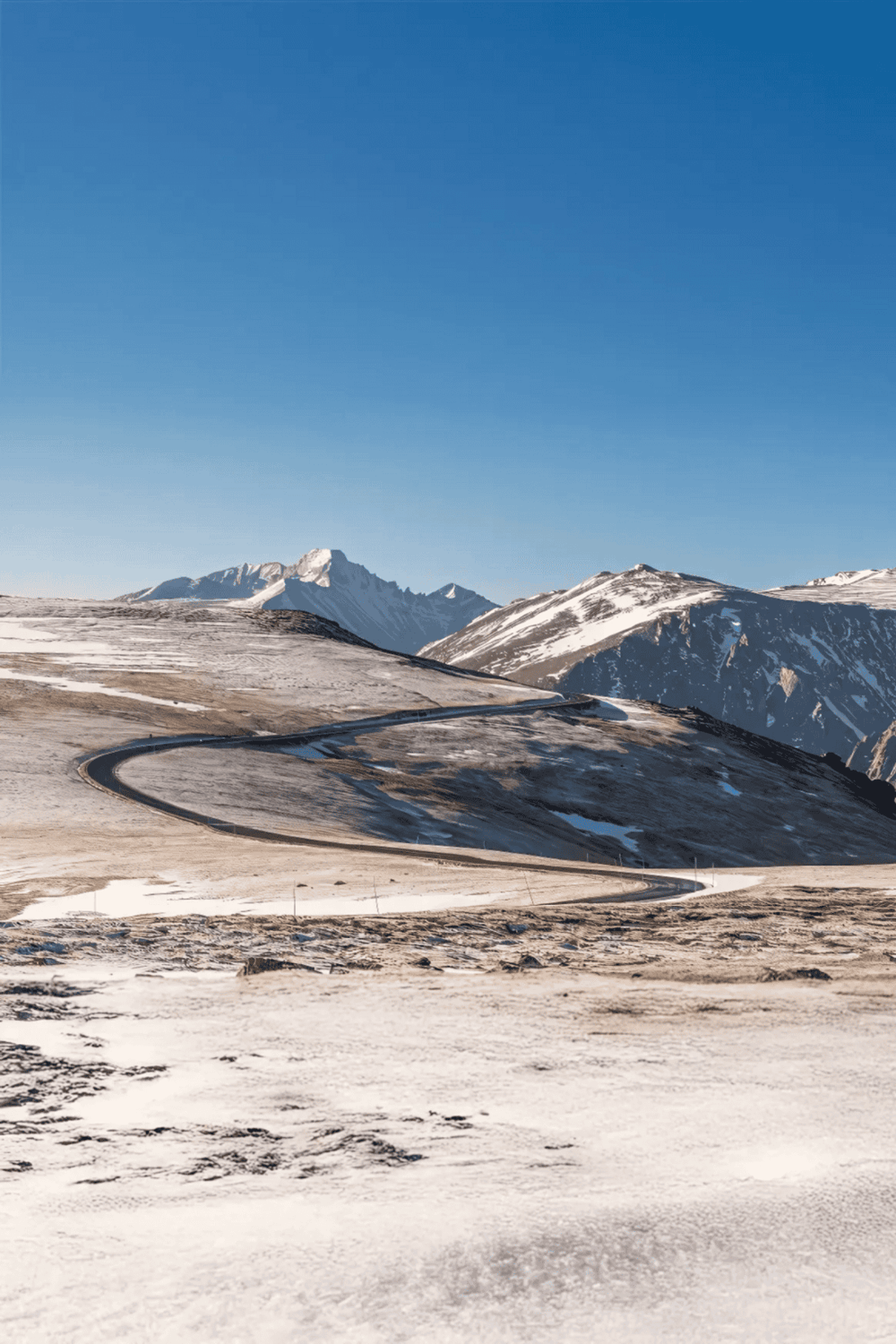 Snow-covered mountain road with winding path, scenic landscape, and clear blue sky.