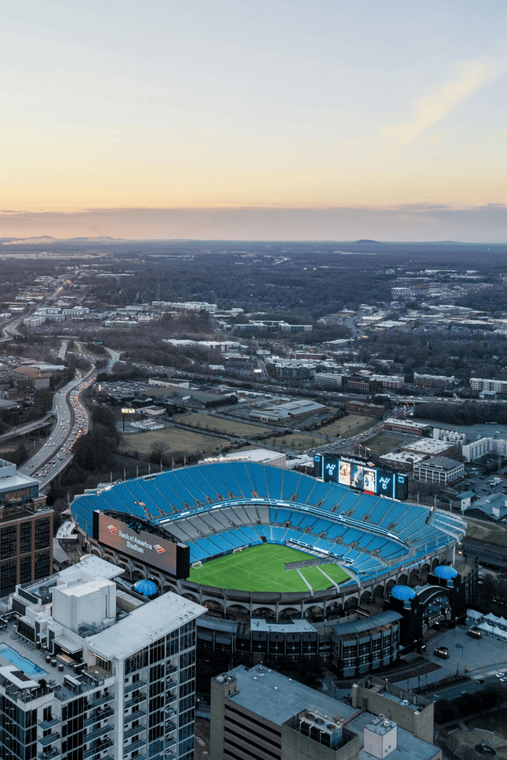 Aerial view of a sports stadium with bright blue seats at sunset, cityscape and highways in background.
