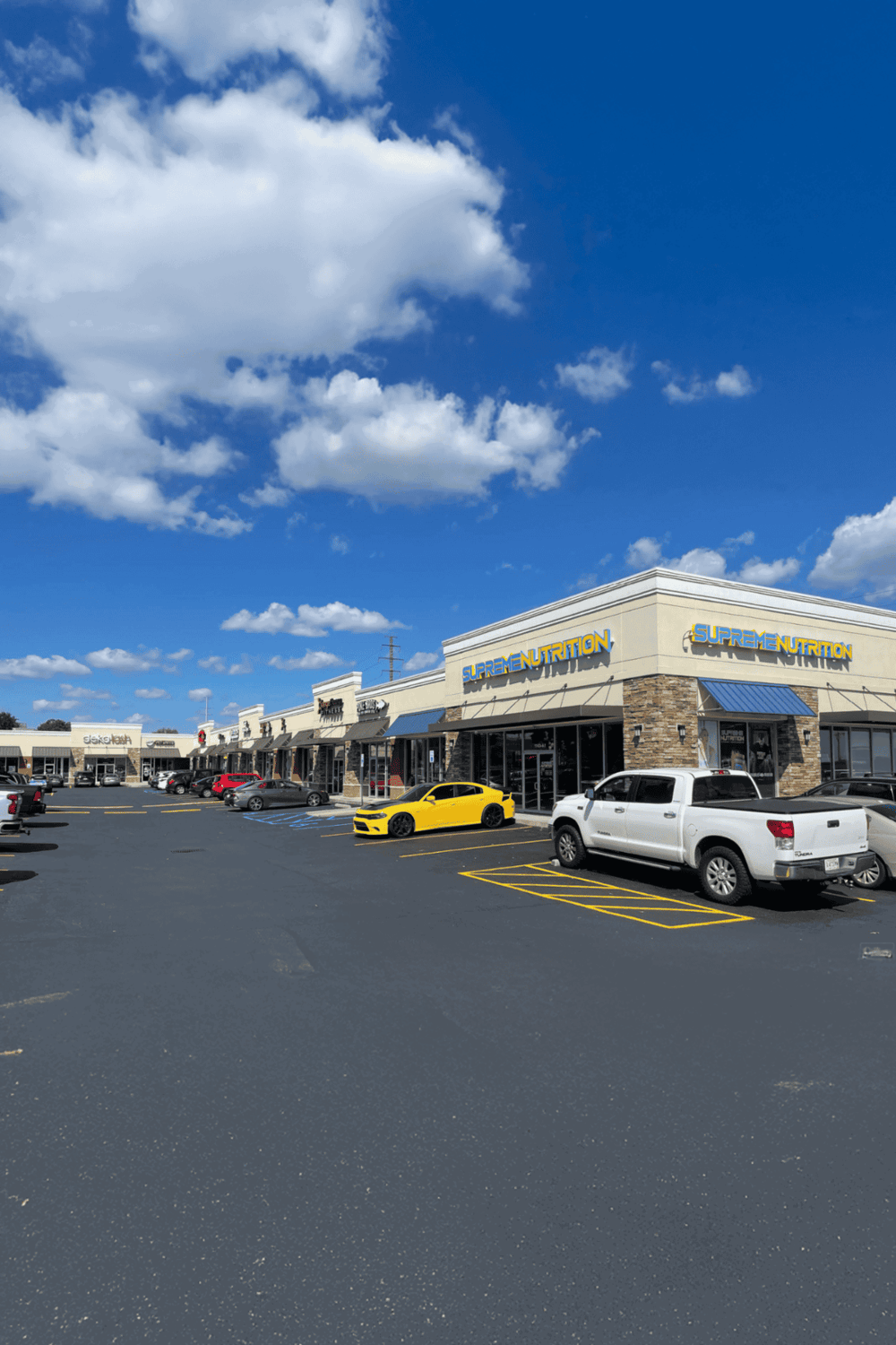 Modern retail shopping plaza with fitness and nutrition stores, clear parking lot, under blue skies.
