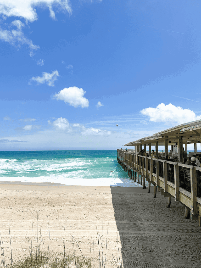 Vibrant beach scene with pier, ocean waves, blue sky, and fluffy clouds.