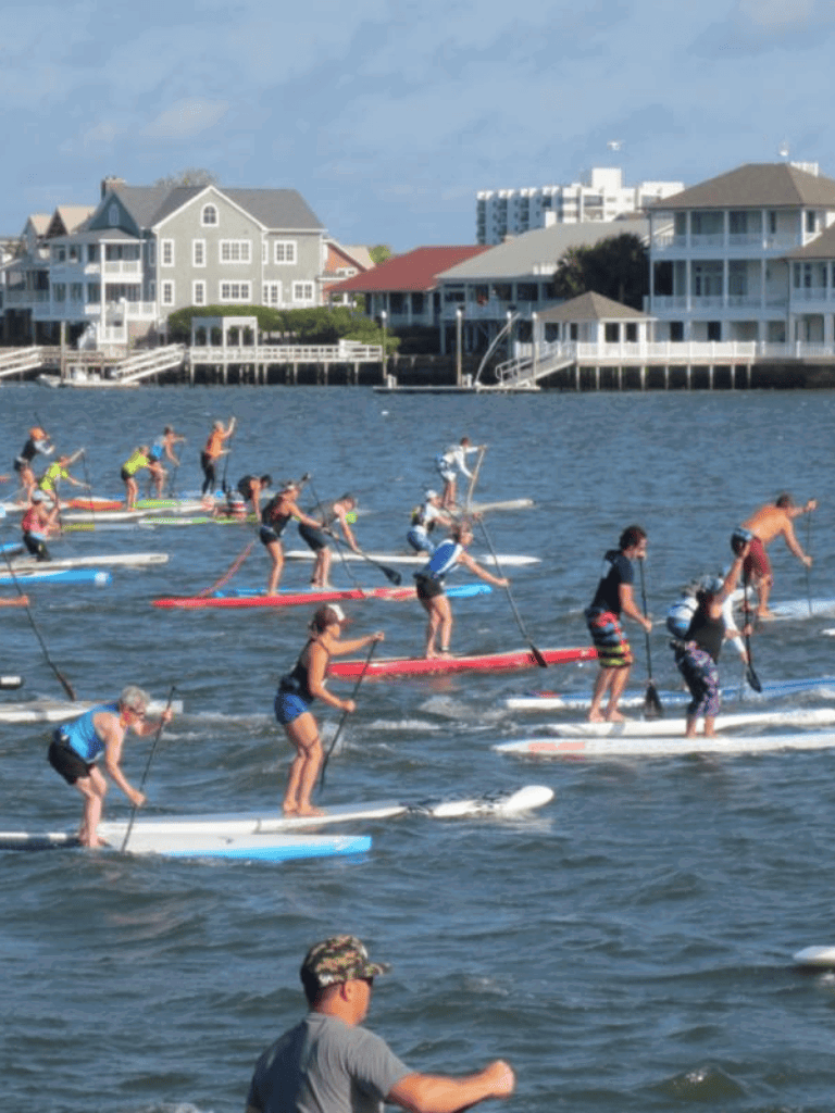 Stand-up paddleboarding contest in waterfront area with houses and buildings in background.