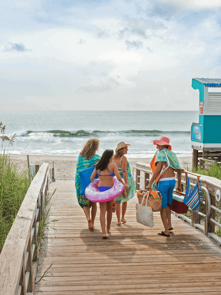 Relaxed beachgoers walking on wooden pier with beach and ocean view, carrying umbrellas and floaties, sunny weather.