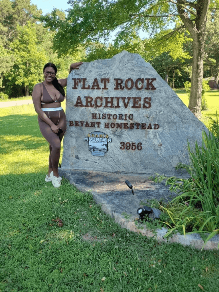 Bright woman in athletic wear stands next to Flat Rock Archives historic landmark in Bryant Homestead, Alabama.