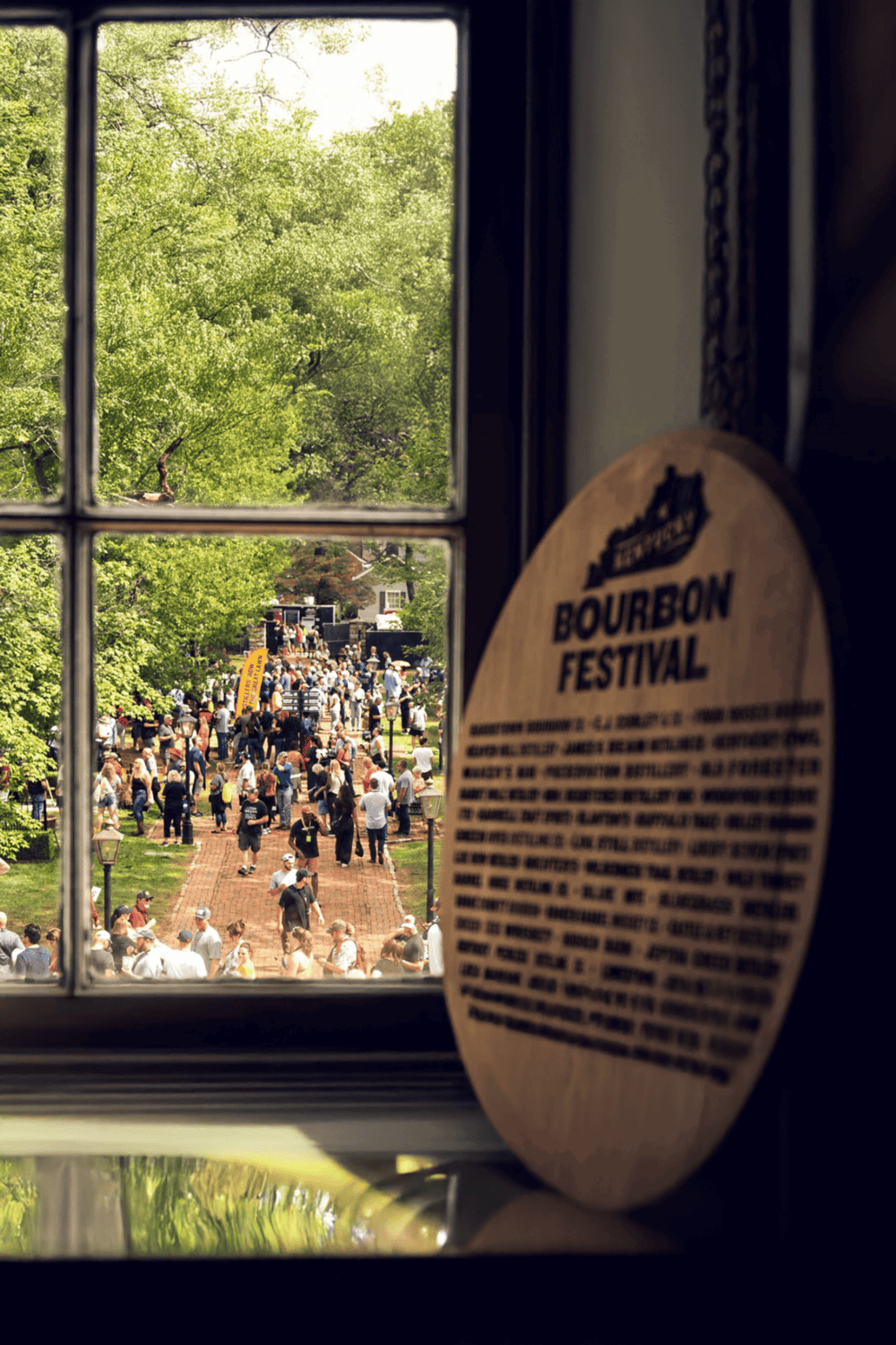 Bourbon Festival crowd seen through window, lively outdoor event with music, food, and vendors.