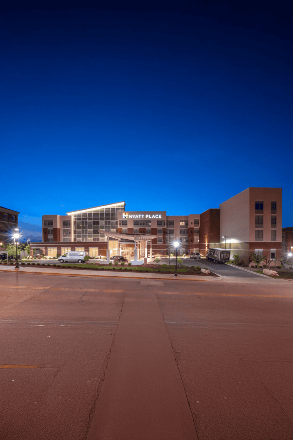Modern Hyatt Place hotel exterior at dusk with illuminated signage and parking area, showcasing hospitality and travel accommodations.