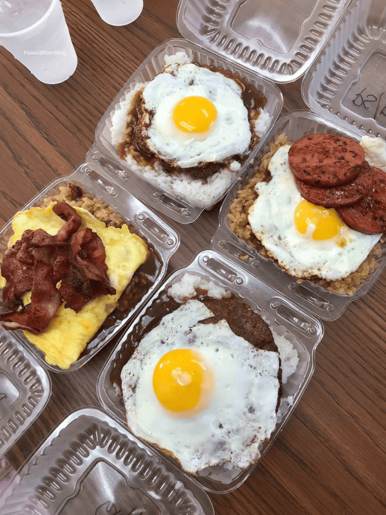 Savory breakfast dishes with fried eggs, rice, bacon, sausage, and breakfast meats in takeout containers.
