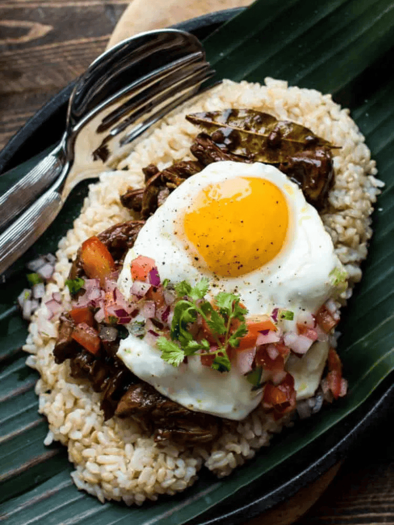 Savory rice bowl with crispy egg, seasoned beef, and fresh pico de gallo on banana leaf.