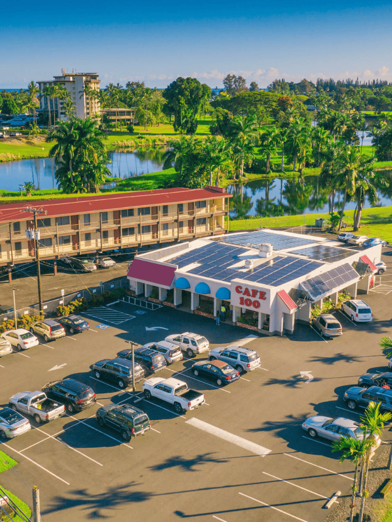 Modern shopping plaza with Cafe 100 and surrounding parking lot near lush tropical landscape.